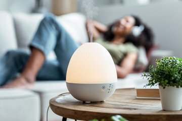 Essential oil aroma diffuser humidifier diffusing water articles in the air while woman listening music lying on coach. - obrazy, fototapety, plakaty