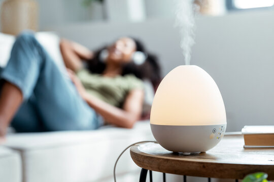 Essential oil aroma diffuser humidifier diffusing water articles in the air while woman listening music lying on coach.