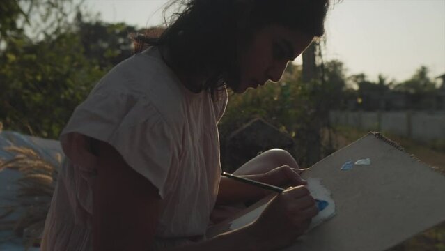 Slow motion panning shot of a talented indian artist sitting in nature while painting an abstract painting with brush and acrylic paint at a beautiful sunset