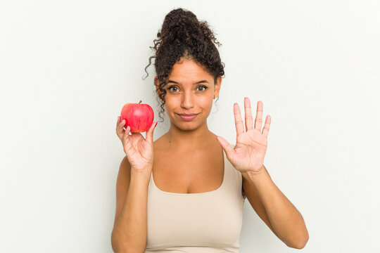 Young brazilian woman holding a red apple isolated smiling cheerful showing number five with fingers.