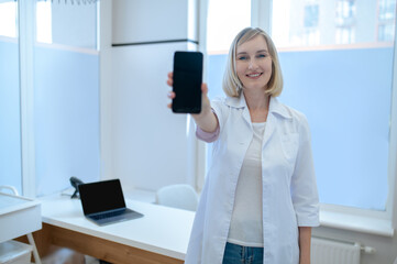 Joyous healthcare worker with the cellphone standing in her office