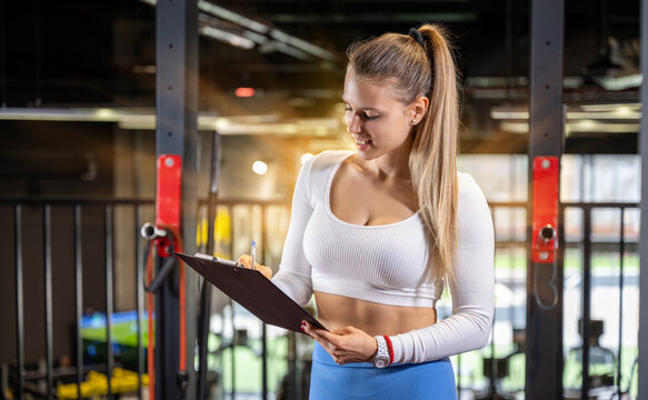 Young fitness instructor making schedule for a client on the clipboard at the gym. Attractive blonde trainer writing report for a client.