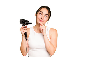 Young caucasian woman holding a hairdryer on green chroma background looking sideways with doubtful and skeptical expression.