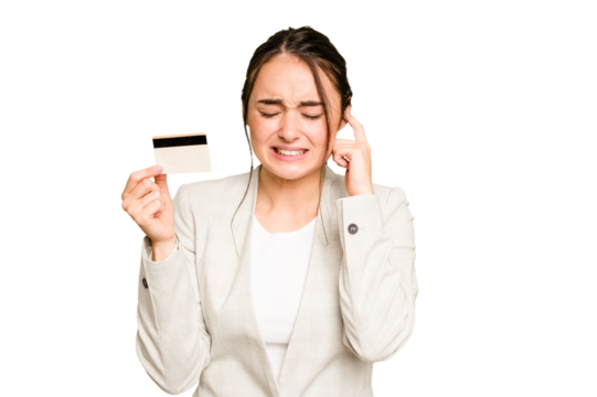 Young caucasian woman holding a credit card isolated on green chroma background covering ears with hands.
