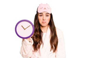 Young caucasian woman holding a clock wearing a pajama isolated on green chroma background shrugs shoulders and open eyes confused.