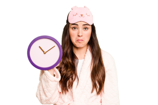 Young caucasian woman holding a clock wearing a pajama isolated on green chroma background shrugs shoulders and open eyes confused.
