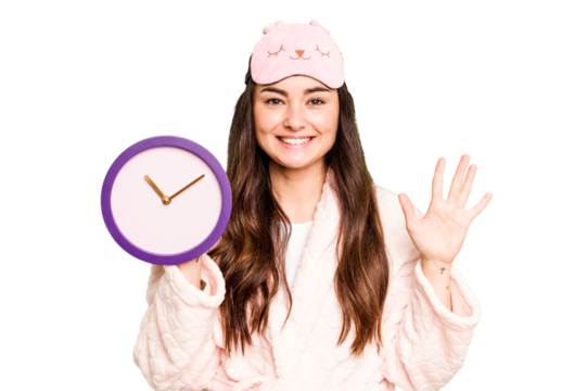 Young caucasian woman holding a clock wearing a pajama isolated on green chroma background smiling cheerful showing number five with fingers.