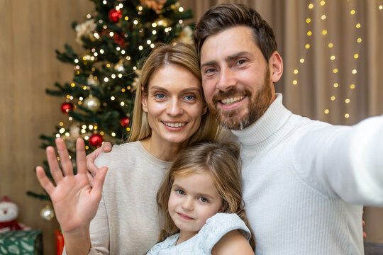 Festive portrait of a young happy family near the Christmas tree at home. Mom, dad and daughter take a selfie, look at the camera, smile, wave.