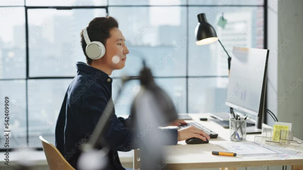 Poster portrait of an asian man smiling while using computer and headphones in bright modern office. commun