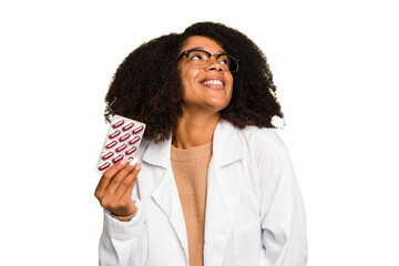 Young pharmacist African American woman holding a tablet of pills isolated dreaming of achieving...