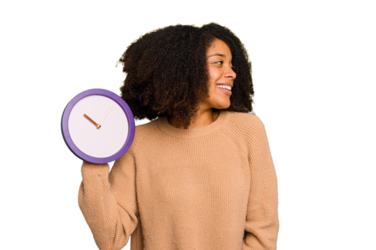Young African American holding a clock isolated looks aside smiling, cheerful and pleasant.