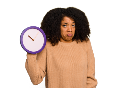 Young African American holding a clock isolated shrugs shoulders and open eyes confused.