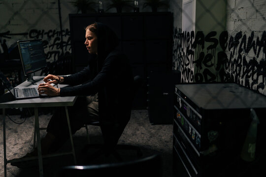 Wide shot of confident hacker man in black sweatshirt using laptop and desktop computer to hack network system from dark room. Concept of cyber attack, virus, malware, illegally and cyber security.