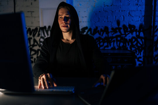 Portrait of hidden hacker man wearing sweatshirt with hood engaged in hacking into security systems, sitting in dark basement room with blue neon lights. Network hackers steal computer system data