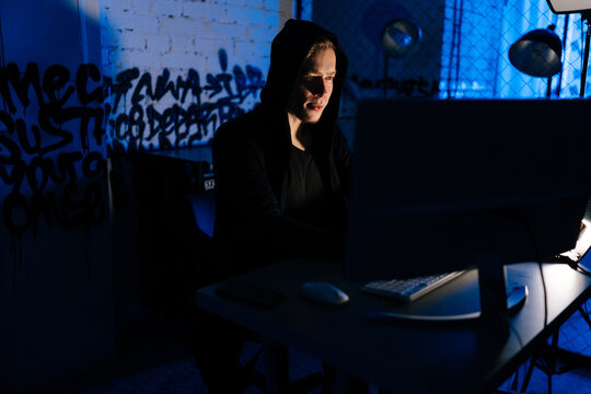 Medium shot portrait of hidden hacker man wearing sweatshirt with hood engaged in hacking into security systems in dark basement room with blue neon lights. Concept of cyber attack, virus, malware.