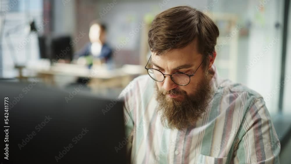 Poster Portrait of Young Caucasian Bearded Man with Glasses Focused and Working on Computer in a Bright Spacious Office. Male Administrator Smiling after Successfully Finishing a Task. Slow Motion Close Up