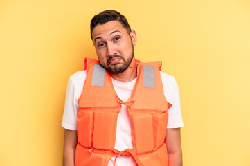 Young latin beach watcher wearing a lifejacket isolated shrugs shoulders and open eyes confused.