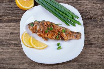 Fried breaded fish with green onions and lemon, in a plate on an old wooden background