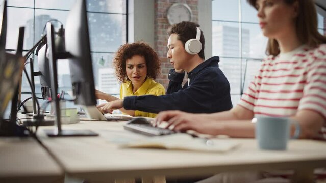 Group of Colleagues Working On Computers in Office. Female Biracial Help Desk Coordinator Collaborates with Asian Male Customer Service Agent with headophones, They Discuss Project, Chat and Smile