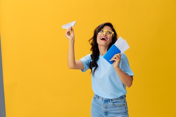 Traveler woman with yellow suitcase, passport and ticket in hand, paper plane, blue t-shirt and jeans on yellow background tourist, travel happiness, glasses, copy space