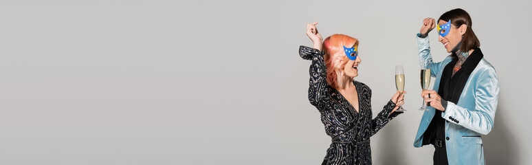 excited and elegant queer friends with champagne glasses dancing at christmas party on grey background, banner