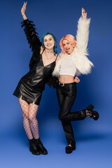 full length of nonbinary person sticking out tongue while posing with happy friend on blue background