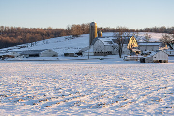 Amish farm in the winter at the base of a snowy hill with an empty field in front on a clear sunny...