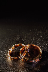 Obraz na płótnie Canvas Gold wedding rings on black background with drops in a strip of light. Wedding attributes.Wedding preparation.
