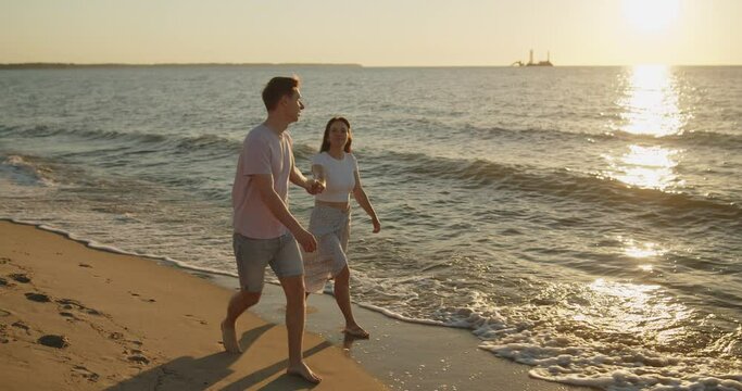 Young loving couple spend time on the beach at sunset. Man and woman enjoying relaxing walk on the beach holding hands. Travel vacation lifestyle.