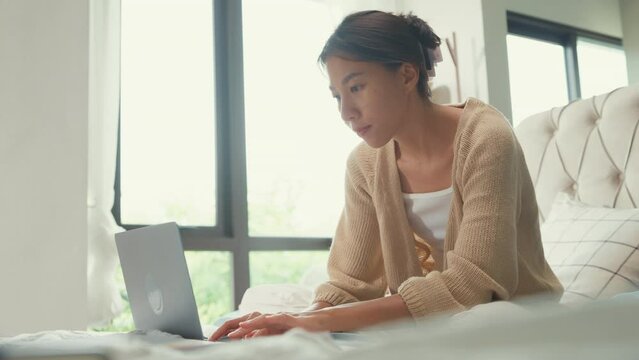 Happy young Asian girl with white cream cardigan on bed focus computer laptop full of paperwork messy document work idea in cozy bedroom at home in morning. Stay quarantine, Work from home concept.