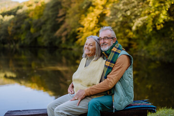 Senior couple in love sitting together on bench near lake, during autumn day.