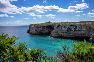 Fototapeta premium Cliffs of Mallorca in summer with a blue sky and clouds. In the foreground are green bushes. In the distance the open blue sea with gray cliffs. vacation, landscape, journey
