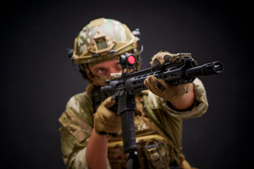 Fototapeta premium Military man in camouflage holding assault rifle target acquisition through monocular. Special forces man dressed up in military ammunition with gun isolated on black background aiming for a shot