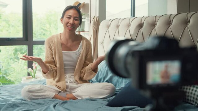 Happy Asian girl influencer with white cream pajamas recording sharing morning routine in camera sit on fluffy comfort bed talk with subscriber in bedroom morning light. Female morning vibes concept.