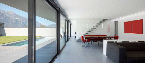 Front view of a modern living room with a dark sofa, behind it a table with chairs and the cantilevered staircase. Outside the window is the green parto and swimming pool