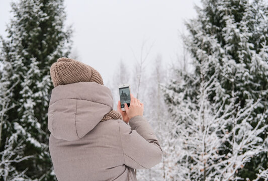 Young woman taking pictures of winter forest using phone