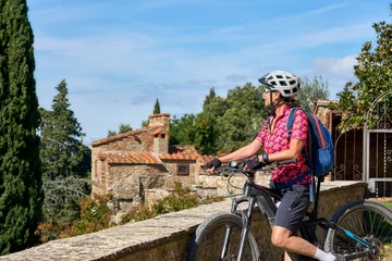 Foto auf Glas cheerful senior woman cycling with her electric mountain bike in a typical Tuscan village near Pienza, Chianti area,Tuscany, Italy © Uwe