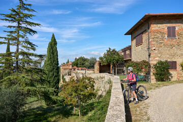 cheerful senior woman cycling with her electric mountain bike in a typical Tuscan village near...