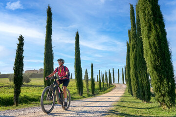 Fototapeta na wymiar nice senior woman riding her electric mountain bike in a cypress avenue in the Chianti area near Pienza, Tuscany , Italy