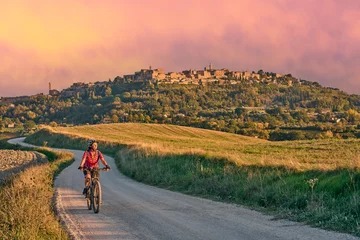 Fotobehang nice senior woman riding her electric mountain bike between olive trees in the Ghianti area with medieval city of Montepulciano in background, Tuscany , Italy © Uwe