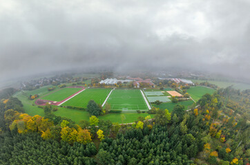 Aerial of a soccer pitch in the fall season with a lot of fog and a forest in the foreground