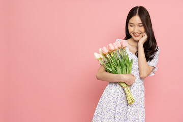 Pretty smiling asian woman wearing a dress is holding a bouquet of flowers isolated on pink...