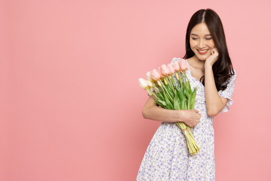 Pretty smiling asian woman wearing a dress is holding a bouquet of flowers isolated on pink background
