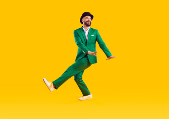 Fototapeta na wymiar Specifics of Irish culture. Cheerful man in green modern suit and hat is dancing and having fun celebrating St. Patrick's Day. Full length festive young man on orange background. Idea for banner.