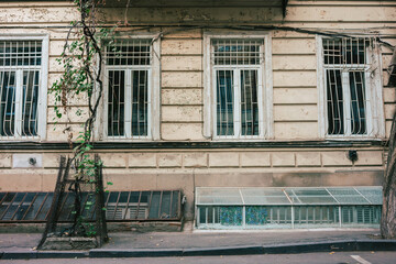 House facade of an old, beige residential building in the old town of Tbilisi. Four barred white windows, a weathered house wall and a sidewalk in the foreground. City trip, travel, tourism, Georgia