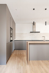 Kitchen with long white marble countertops with one wall covered with plain gray designer cabinets and a column of appliances, island with gray cabinets and wooden countertop
