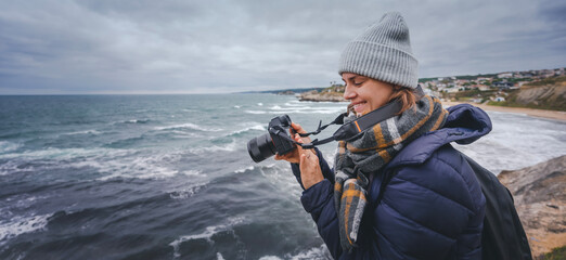 Obraz premium A young cheerful woman in a scarf jacket and a hat, a traveler professional photographer with a camera in her hands on the shore of the winter sea