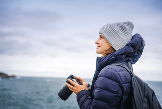 A young cheerful woman in a scarf jacket and a hat, a traveler professional photographer with a camera in her hands on the shore of the winter sea