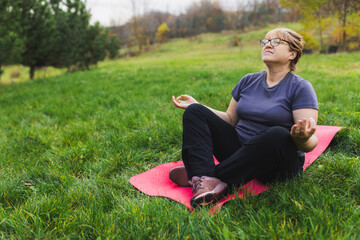 Mature caucasian retired woman in the park meditates and performs breathing exercises, summer day