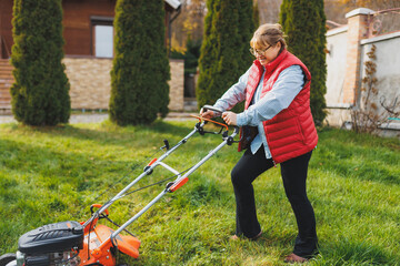 Middle aged woman in red vest using lawn mower on backyard, looking at camera. Female gardener working in summer or autumn, cutting grass in backyard. Concept of gardening, work, nature.